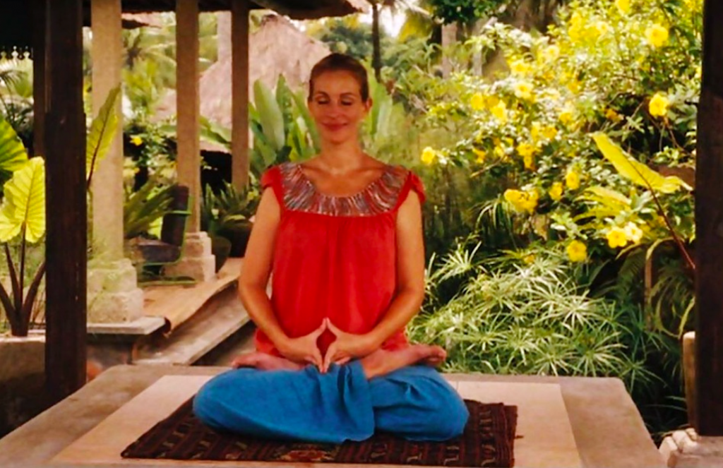 Elizabeth Gilbert, the protagonist, meditates serenely at a retreat in Bali. Dressed in a red blouse and blue pants, she is surrounded by lush tropical vegetation and yellow flowers, symbolizing the balance and spiritual connection she seeks during this stage of her journey of self-discovery.